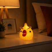 A glowing yellow chick-shaped night lamp with a red beak and black eyes sits on a white nightstand next to a lamp.