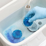 A gloved hand placing a blue toilet cleaner tablet into a toilet tank with blue water.