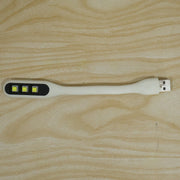 A white flexible USB LED light with three yellow LED bulbs and a USB-A connector resting on a light wood grain surface.