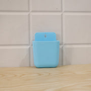 A light blue wall-mounted plastic storage pocket is shown against a white tiled wall and a wooden surface.