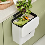 Chopped green vegetables are scraped from a plate into a white wall-mounted compost bin with an open lid.