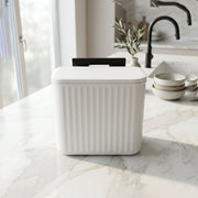 A white ribbed compost bin with a closed lid and black mounting bracket sits on a white marble kitchen counter.