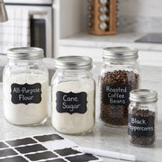Four glass jars with chalkboard labels for flour, sugar, coffee beans, and peppercorns, with blank labels and a white marker.
