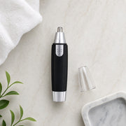 A black and silver nose hair trimmer with its clear cap removed lies on a white marble surface next to a white towel and