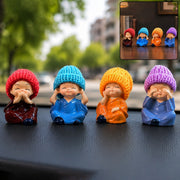 Four resin baby monk figurines with colorful knitted hats sitting on a dark surface outdoors. The first monk wears a red hat