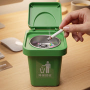 A hand holding a lit cigarette over a green mini trash can ashtray with an open lid on a wooden desk.