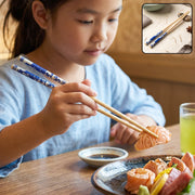 A young girl in a blue shirt uses bamboo chopsticks with blue Chinese print to pick up a piece of salmon.