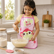 A young girl in a pink monkey apron smiles while whisking batter in a glass bowl in a kitchen.