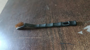 A black-handled brass wire brush rests on a dark brown wooden surface.