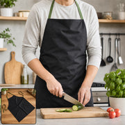 A person in a light grey long-sleeved shirt and a black apron with green straps is slicing a cucumber on a wooden cutting