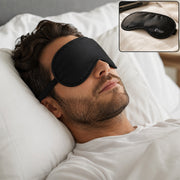 A man with a beard sleeps peacefully on a white pillow, wearing a black sleeping eye mask with an adjustable strap.