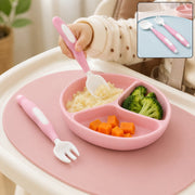 A baby's hand holds a pink and white spoon over a pink divided plate with food, a pink fork rests beside it.