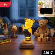 A glowing yellow bear night light and phone stand on a desk, with a small inset showing it holding a phone, and a red 